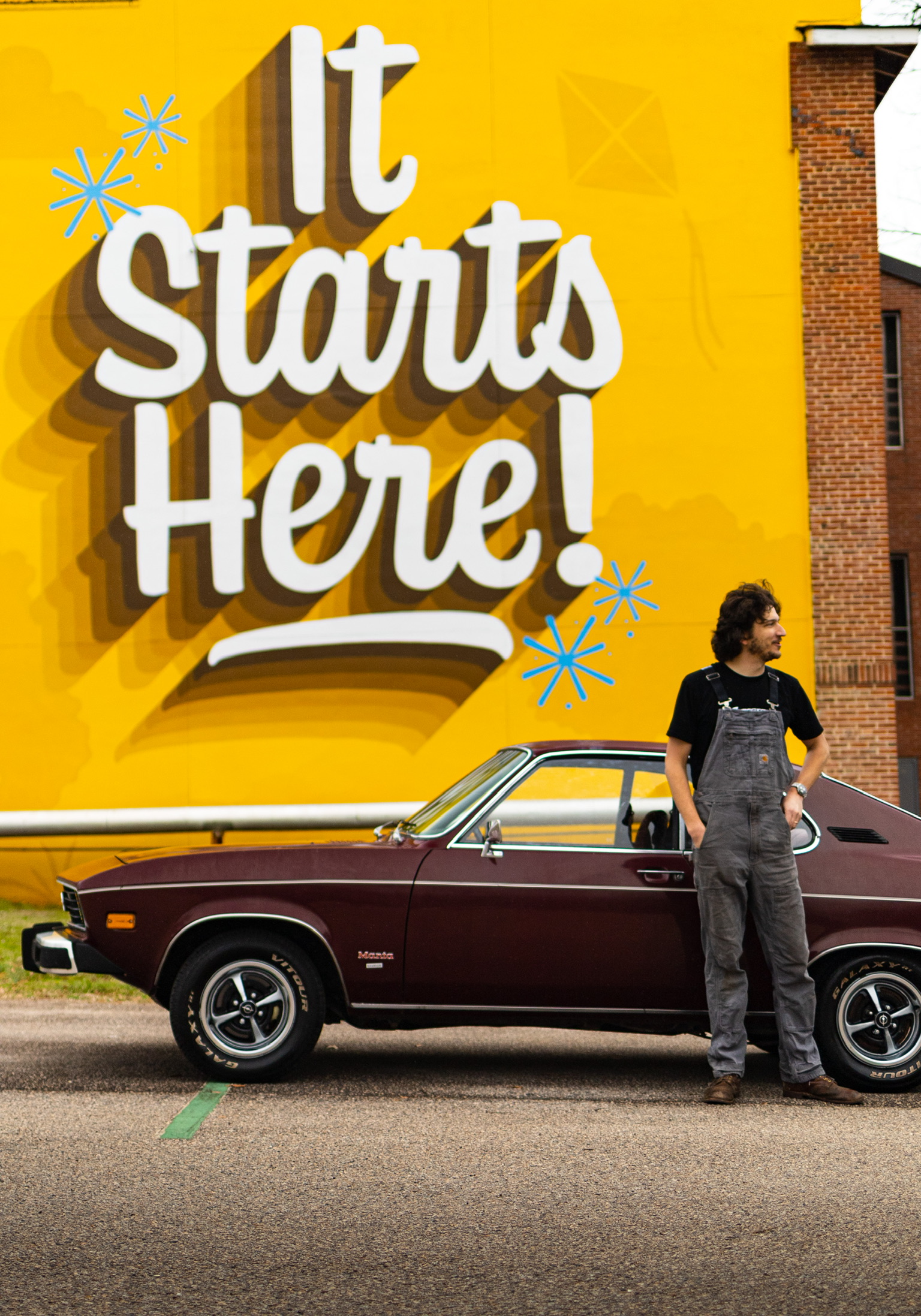 Dustin Simons standing next to his Opel in front of an It Starts Here mural