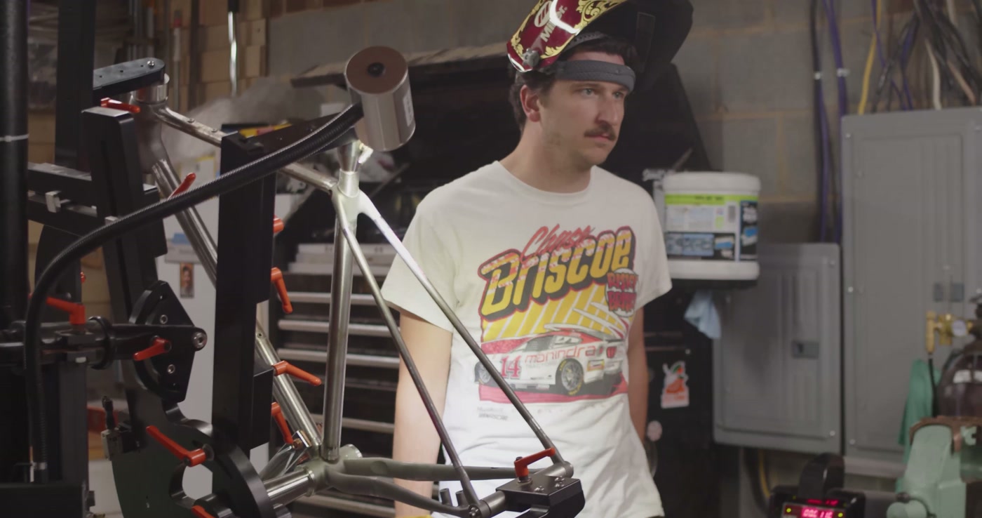 Dustin in the shop with welding helmet raised, frame in the jig